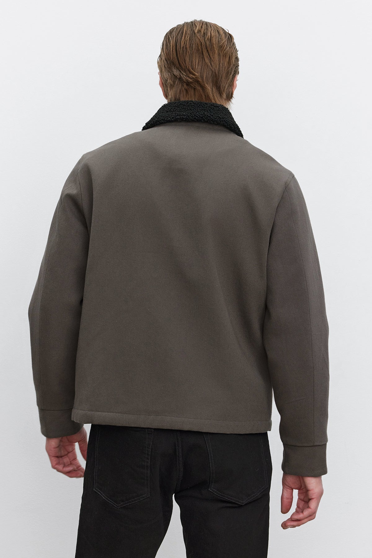   A person with short hair, wearing the Velvet by Graham & Spencer CASH JACKET featuring a dark full-zip design with a black faux sherpa collar and paired with black pants, is seen from the back against a plain background. 