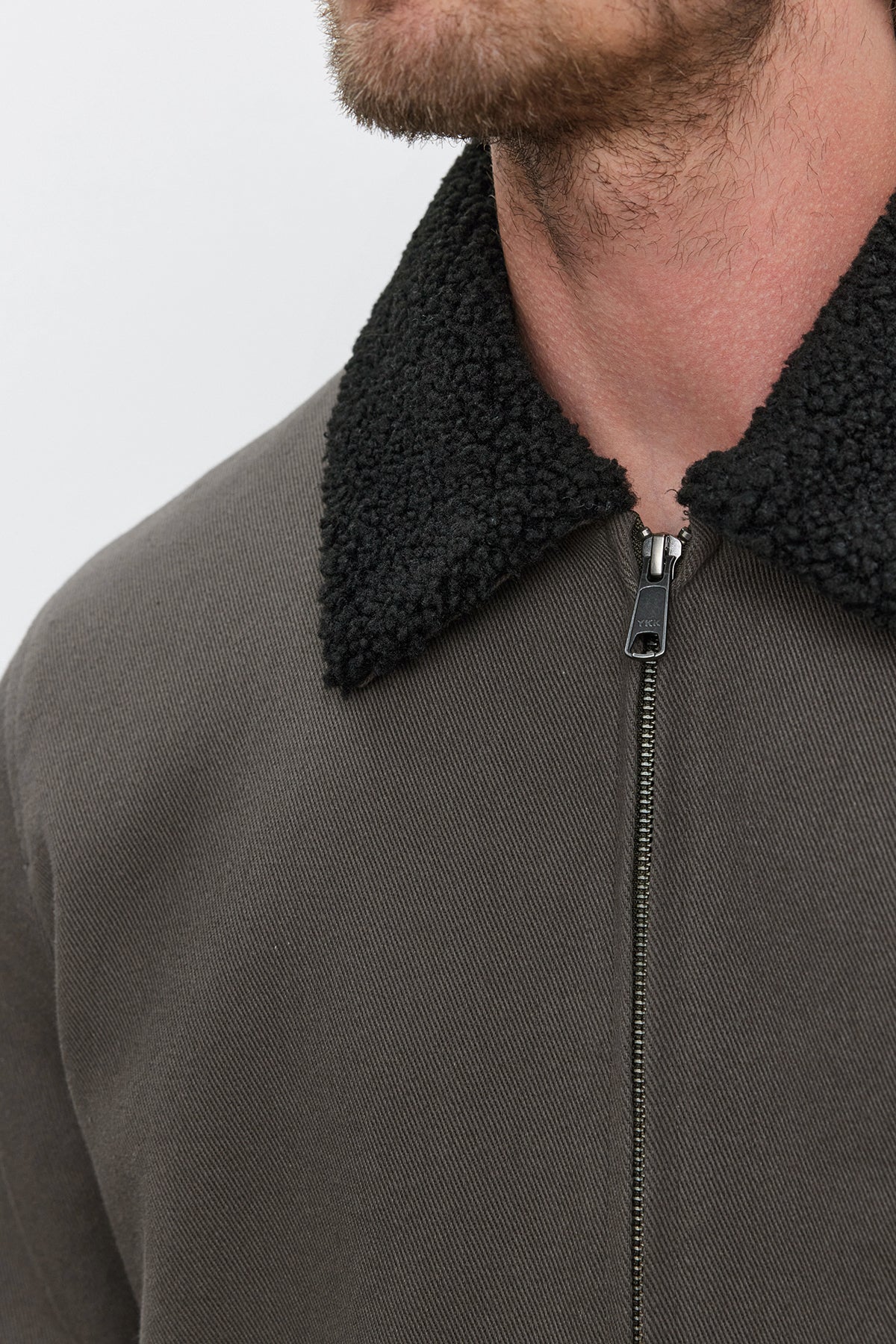   Close-up of a person wearing the Velvet by Graham & Spencer CASH JACKET, which features a dark green full-zip design with a textured black faux sherpa collar. The front is partially zipped, revealing the lower part of the face. 