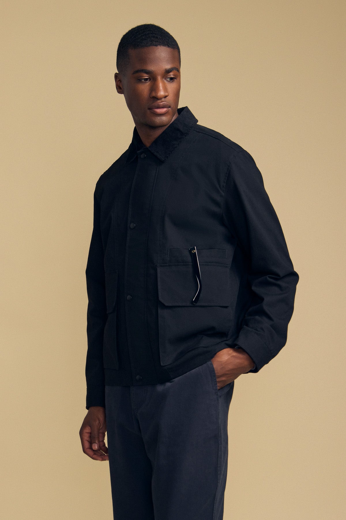 A man stands against a beige background wearing the DESREN JACKET by Velvet by Graham & Spencer—a black cotton canvas jacket featuring large pockets and a corduroy collar—paired with dark pants. He looks to the side with one hand in his pocket.