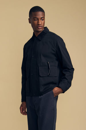 A man stands against a beige background wearing the DESREN JACKET by Velvet by Graham & Spencer—a black cotton canvas jacket featuring large pockets and a corduroy collar—paired with dark pants. He looks to the side with one hand in his pocket.