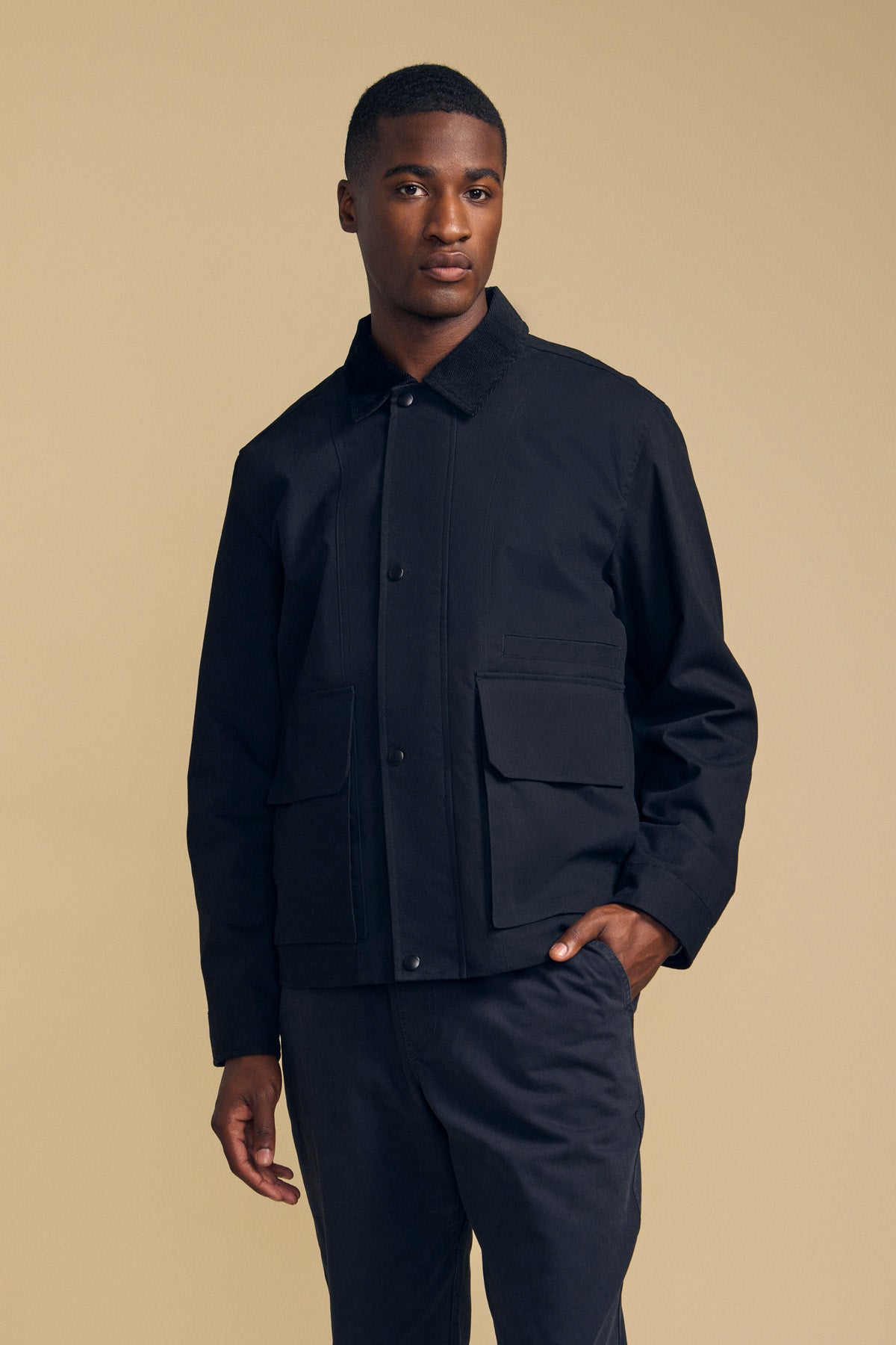 A person stands against a beige background, looking directly at the camera, wearing the DESREN JACKET by Velvet by Graham & Spencer—a black cotton canvas jacket with large front pockets, a subtle corduroy collar for added texture, and matching black pants.