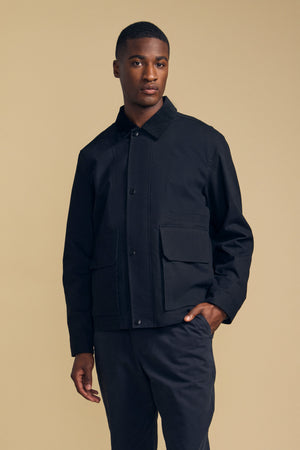A person stands against a beige background, looking directly at the camera, wearing the DESREN JACKET by Velvet by Graham & Spencer—a black cotton canvas jacket with large front pockets, a subtle corduroy collar for added texture, and matching black pants.