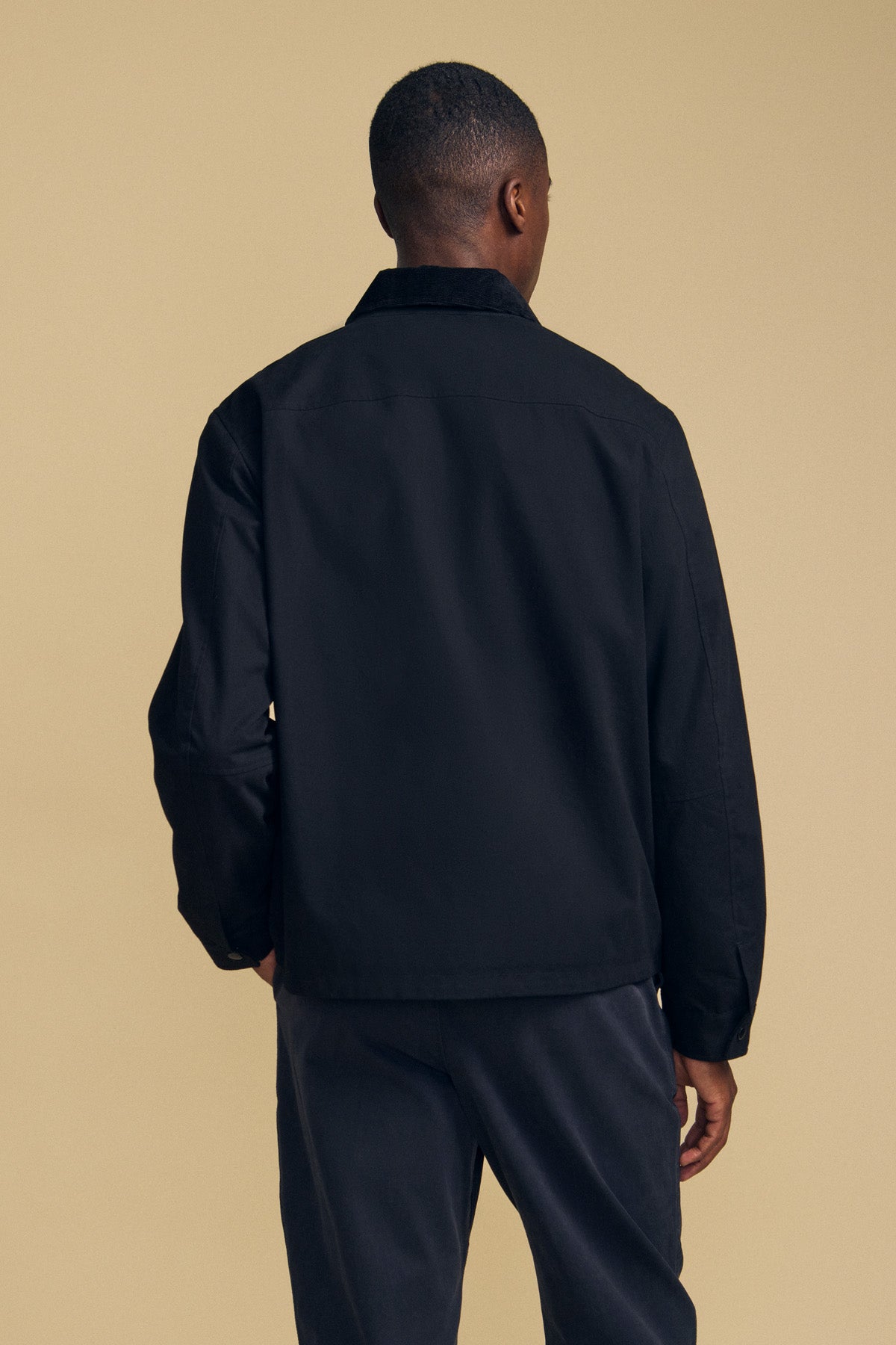A person stands facing away from the camera, wearing the DESREN JACKET by Velvet by Graham & Spencer—a black cotton canvas jacket with a corduroy collar—paired with dark pants against a beige background.