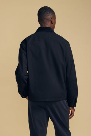 A person stands facing away from the camera, wearing the DESREN JACKET by Velvet by Graham & Spencer—a black cotton canvas jacket with a corduroy collar—paired with dark pants against a beige background.