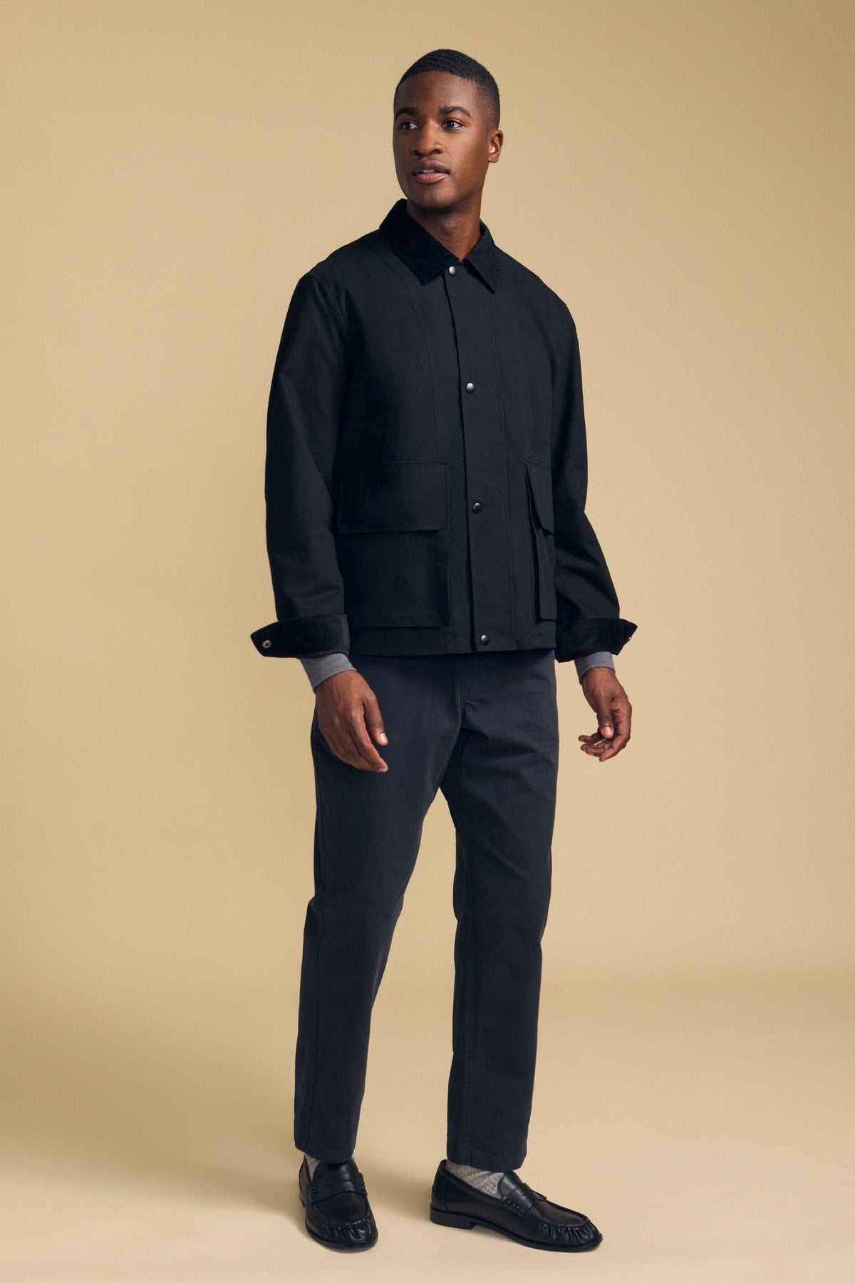 A man stands in a studio against a beige background wearing the DESREN JACKET by Velvet by Graham & Spencer—a black cotton canvas jacket with a subtle plaid lining—paired with matching pants and black loafers.-42092468732097