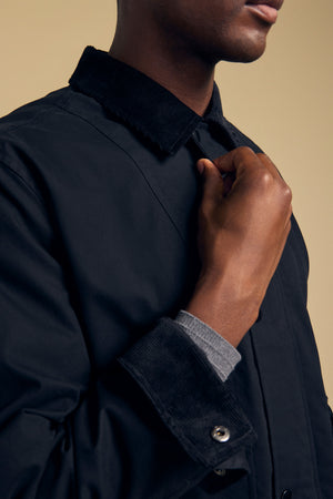 A person adjusts the collar of the Velvet by Graham & Spencer DESREN JACKET, a black cotton canvas jacket with a corduroy collar, worn over a grey long-sleeve shirt.