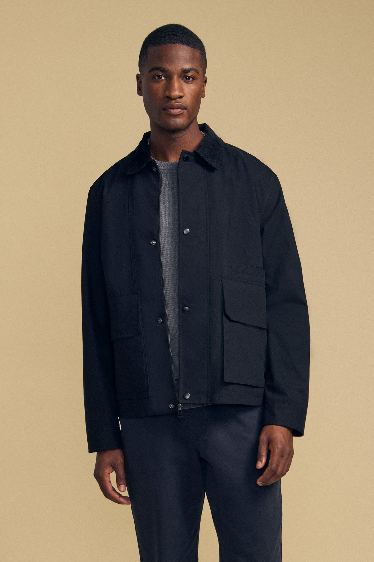 A man stands against a beige background, looking at the camera with a neutral expression, wearing the DESREN JACKET by Velvet by Graham & Spencer—a black cotton canvas jacket with a plaid lining—layered over a gray sweater and paired with dark pants.