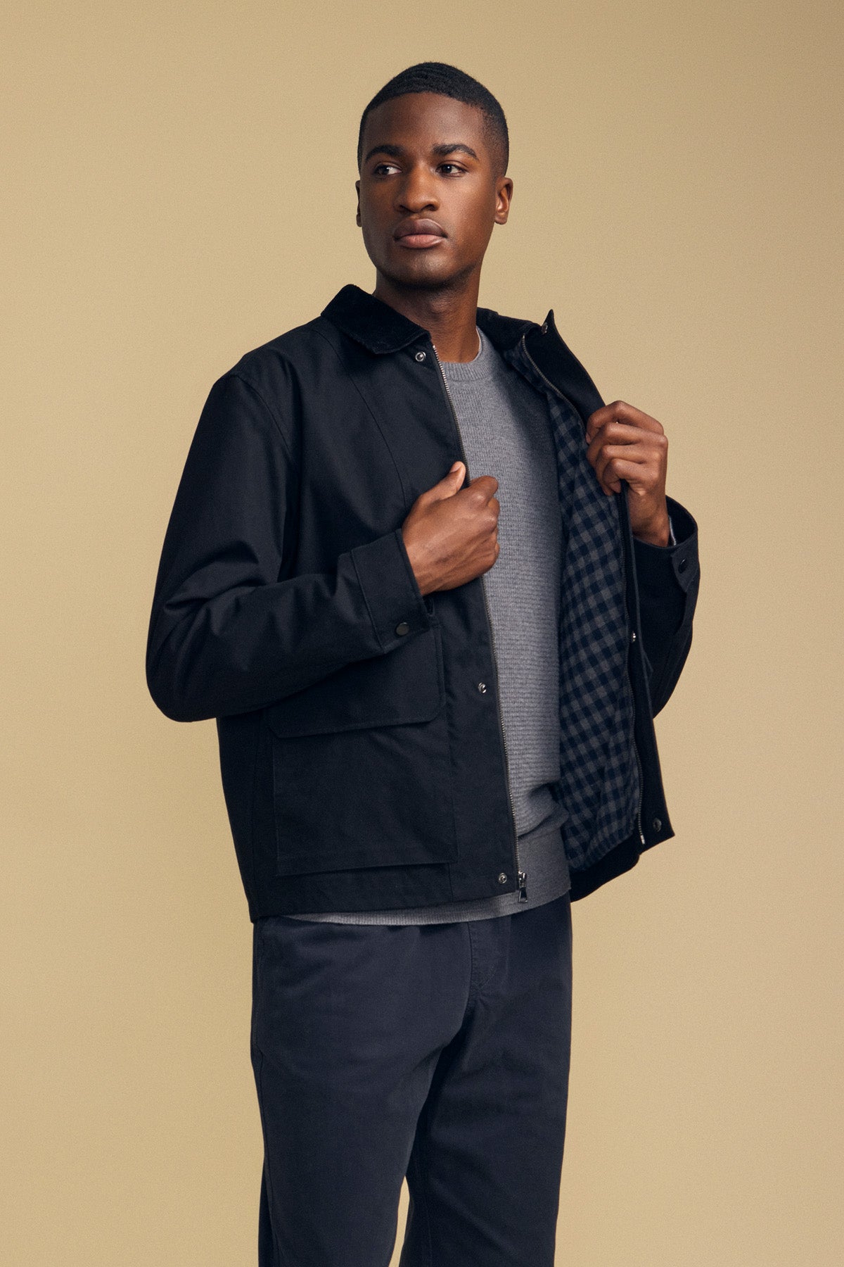 A man stands against a beige background, wearing the DESREN JACKET by Velvet by Graham & Spencer—a black cotton canvas jacket featuring a corduroy collar—layered over a gray shirt and dark pants, holding one side open to reveal its plaid lining.