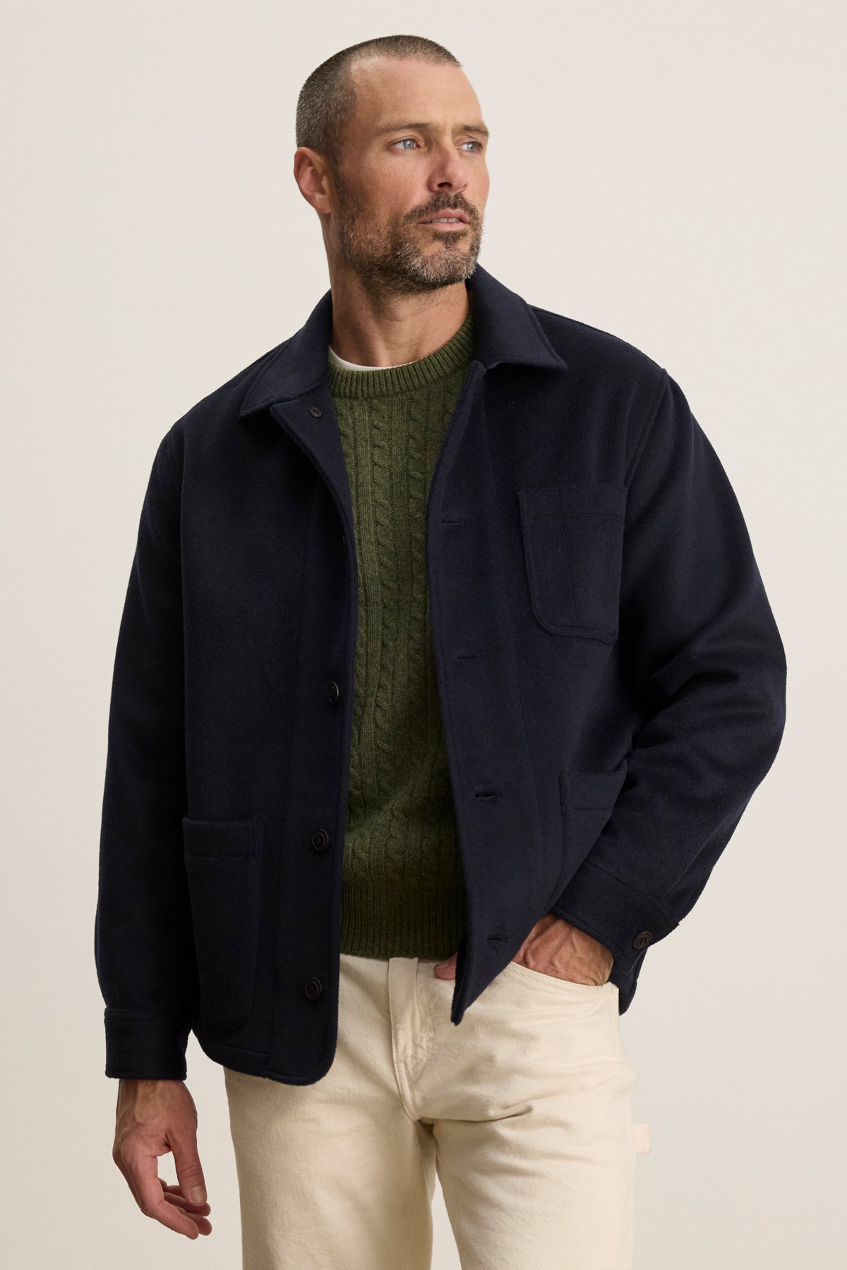 A man with short hair and a trimmed beard wears the Velvet by Graham & Spencer EMERSON CHORE JACKET over a green cable-knit sweater and light pants, standing against a plain light background.