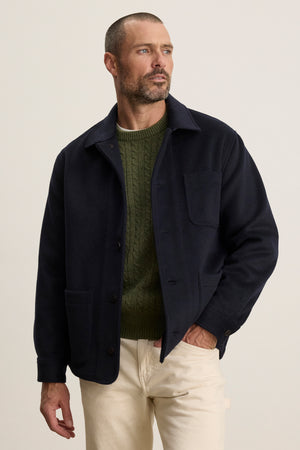 A man with short hair and a trimmed beard wears the Velvet by Graham & Spencer EMERSON CHORE JACKET over a green cable-knit sweater and light pants, standing against a plain light background.