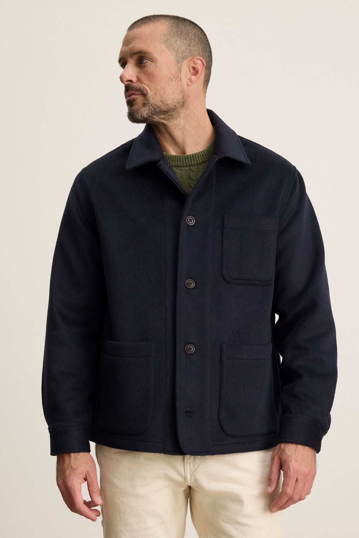 A man with short hair and a beard wears the EMERSON CHORE JACKET by Velvet by Graham & Spencer over a green sweater and light pants, standing against a plain light background.-39934406099137