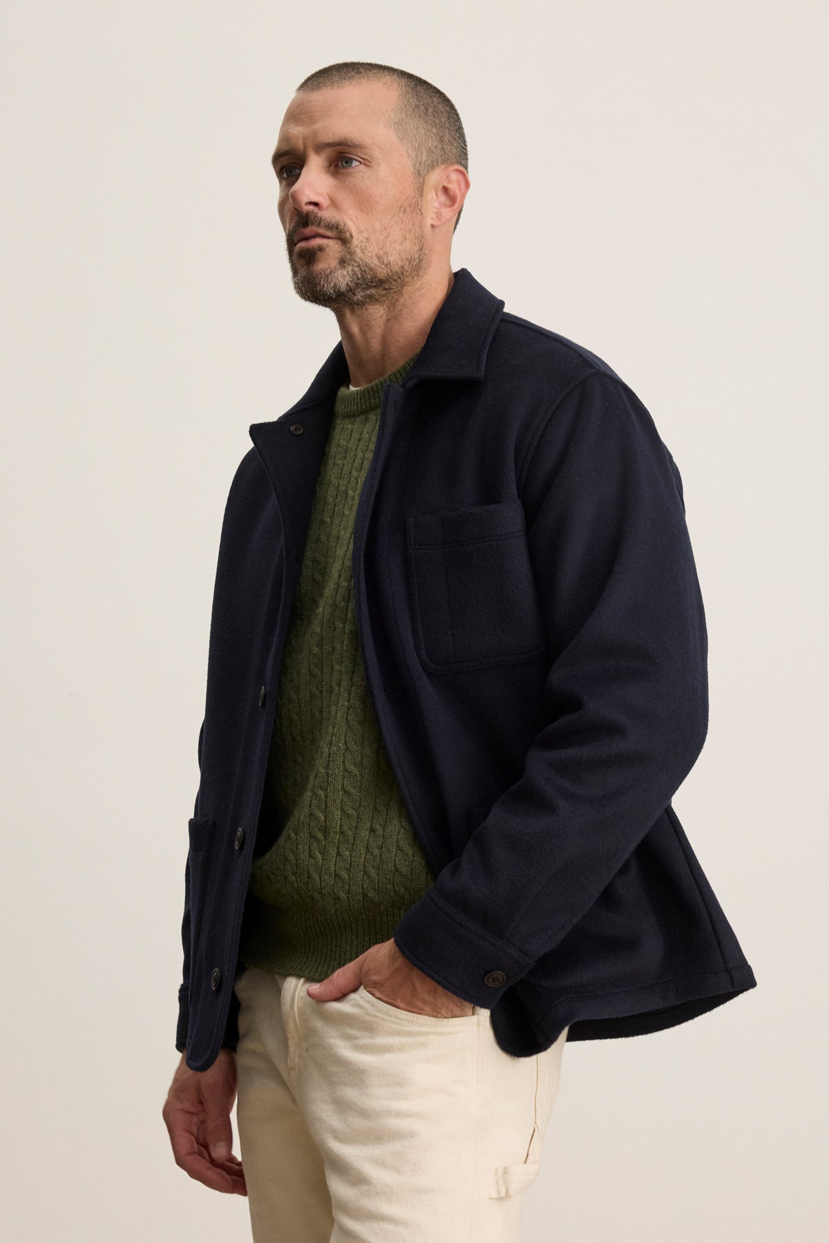 A man with short hair and a beard wears the EMERSON CHORE JACKET by Velvet by Graham & Spencer over a green knit sweater and beige pants, standing against a plain light background with one hand in his pocket.