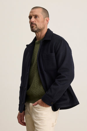 A man with short hair and a beard wears the EMERSON CHORE JACKET by Velvet by Graham & Spencer over a green knit sweater and beige pants, standing against a plain light background with one hand in his pocket.
