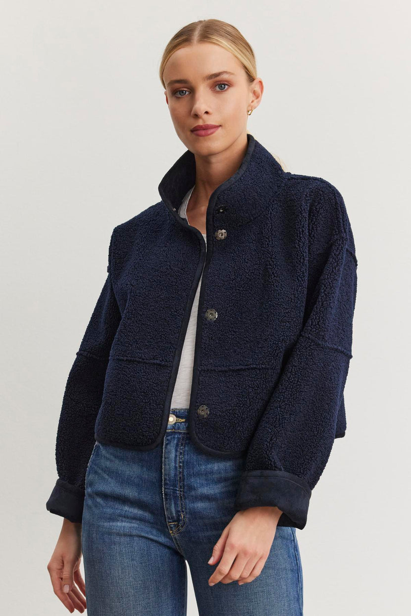 A woman poses against a plain white backdrop, showcasing the Velvet by Graham & Spencer's KELLY LUXE SHERPA REVERSIBLE JACKET in a chic navy blue wool with faux sherpa detailing, layered over a white shirt and blue jeans.