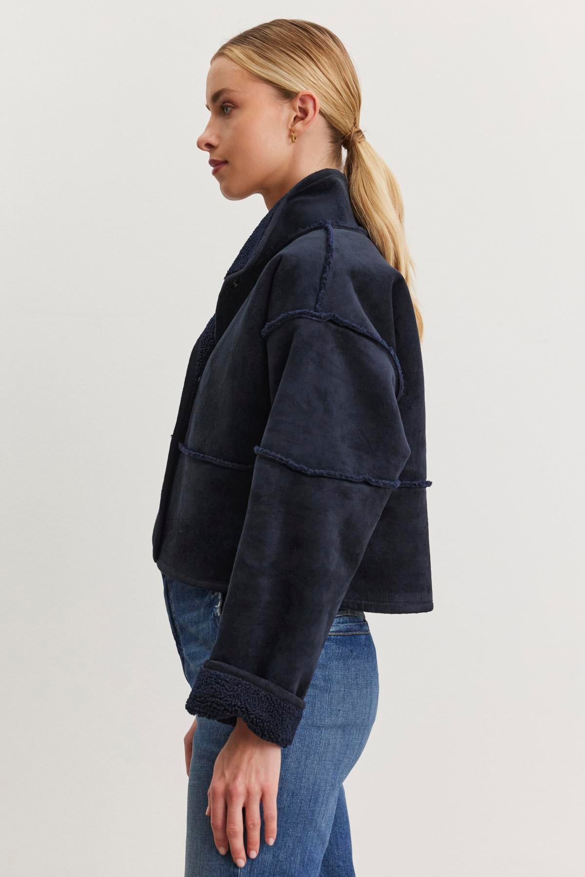   A person wearing the KELLY LUXE SHERPA REVERSIBLE JACKET by Velvet by Graham & Spencer stands sideways in blue jeans, with a ponytail, showcasing its oversized fit, dark color, and high collar. 