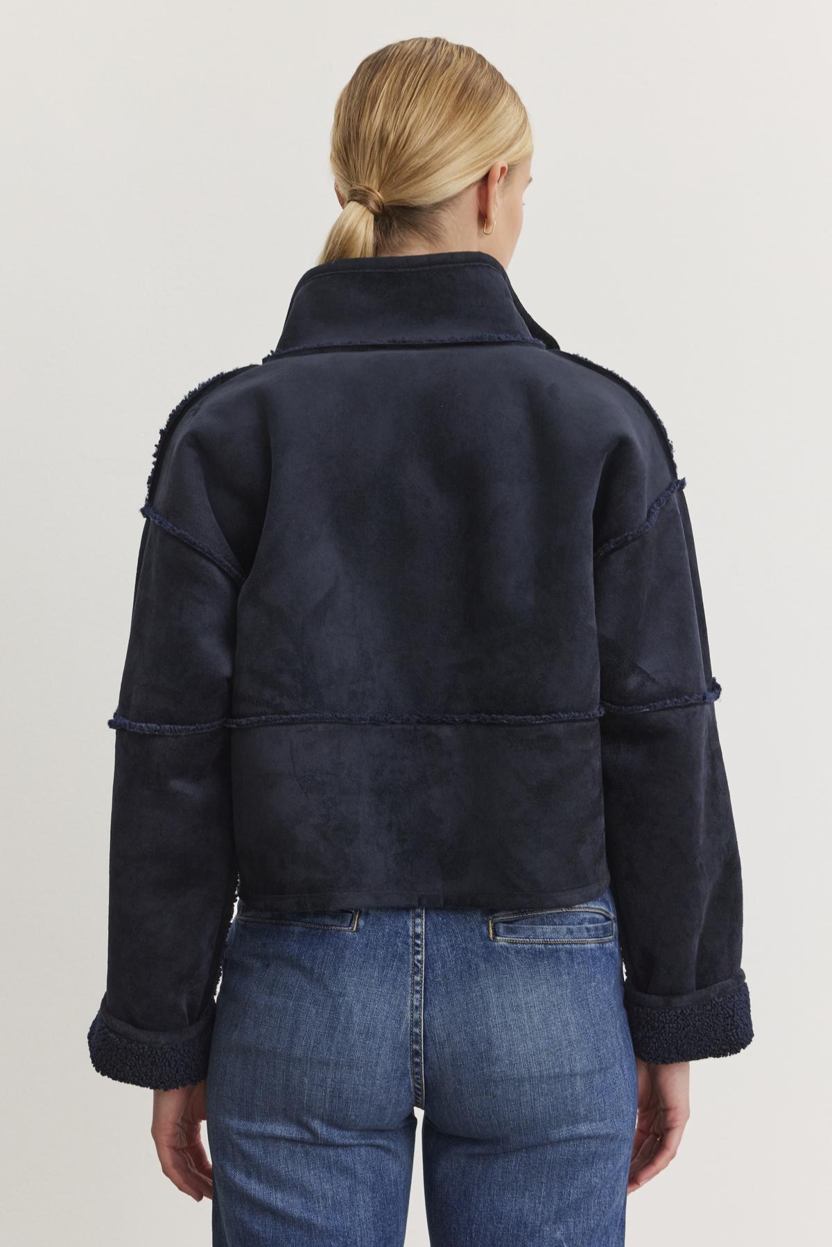   A person with tied-back hair wears the KELLY LUXE SHERPA REVERSIBLE JACKET by Velvet by Graham & Spencer, featuring a high collar and paired with blue jeans, standing with their back to the camera. 