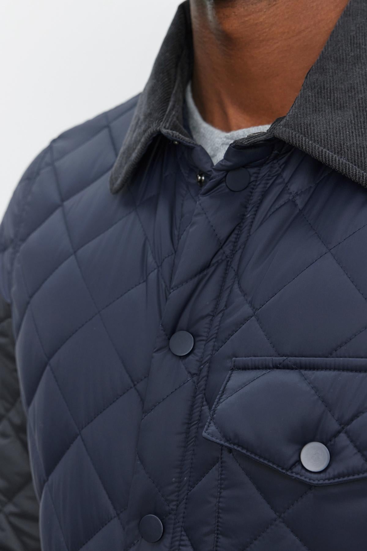   Close-up of a person wearing the LOU JACKET by Velvet by Graham & Spencer, a dark quilted puffer jacket with a button-up front and a contrasting corduroy collar. The jacket also features a small front pocket with a button. 