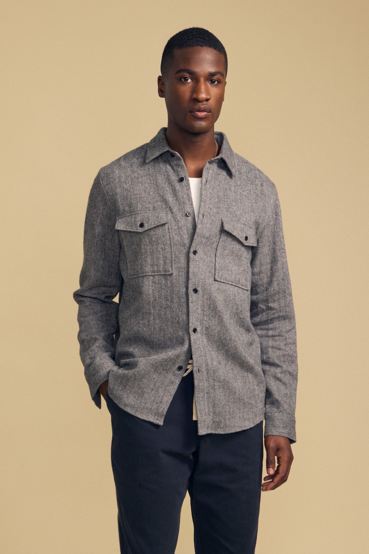 A man stands against a beige background wearing the Velvet by Graham & Spencer MAURICE SHIRT JACKET, a gray button-up shirt with chest pockets, paired with dark pants and looking at the camera.
