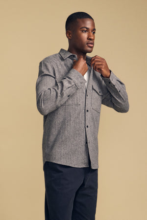 A man stands against a beige background, buttoning the MAURICE SHIRT JACKET by Velvet by Graham & Spencer over a white undershirt and wearing dark pants.