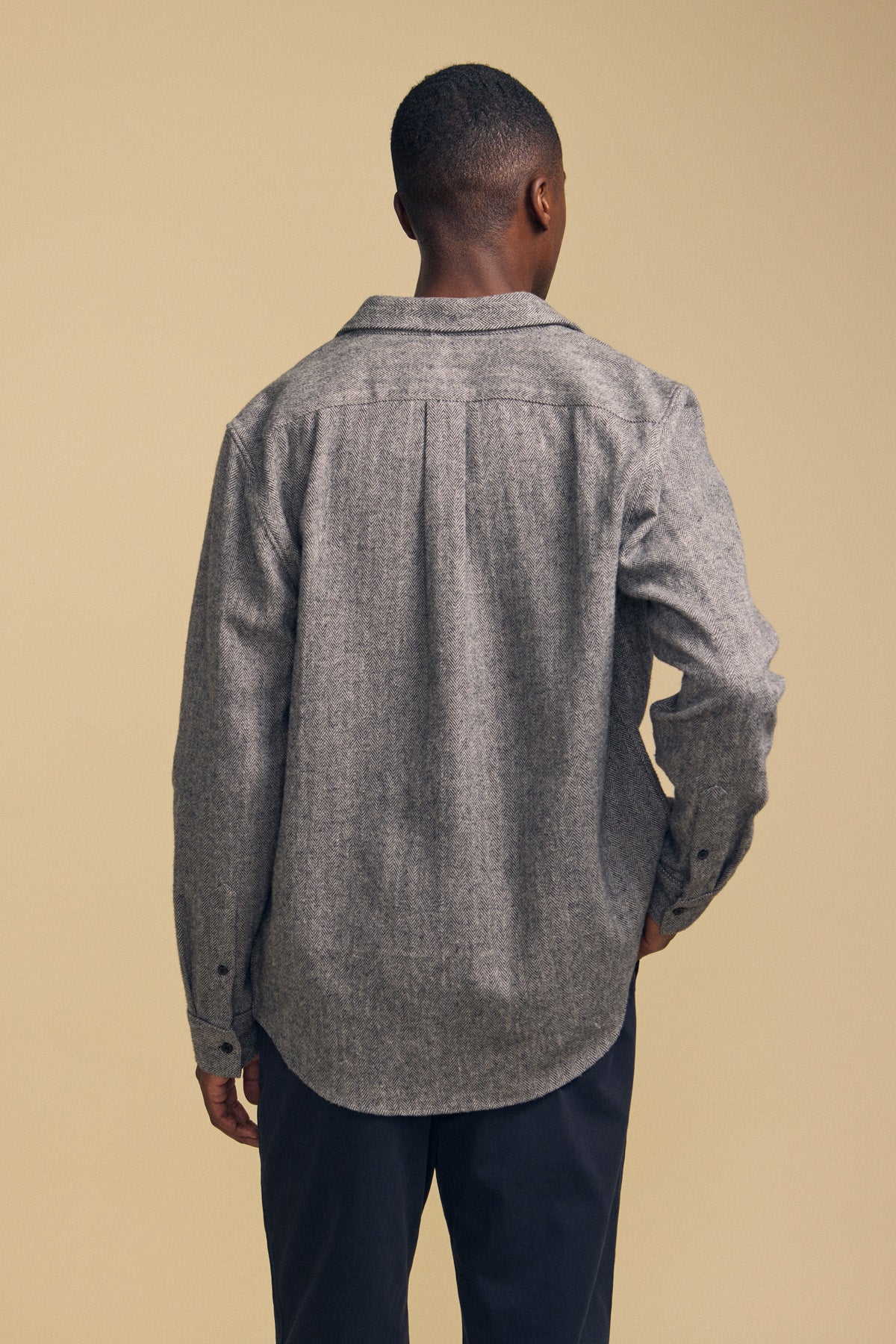 A man stands facing away from the camera, wearing the MAURICE SHIRT JACKET by Velvet by Graham & Spencer in grey, paired with dark pants against a beige background.