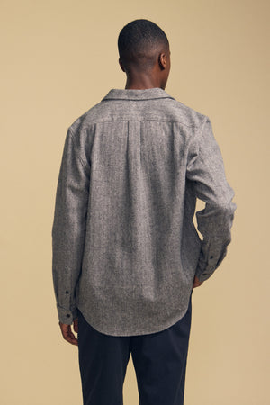 A man stands facing away from the camera, wearing the MAURICE SHIRT JACKET by Velvet by Graham & Spencer in grey, paired with dark pants against a beige background.