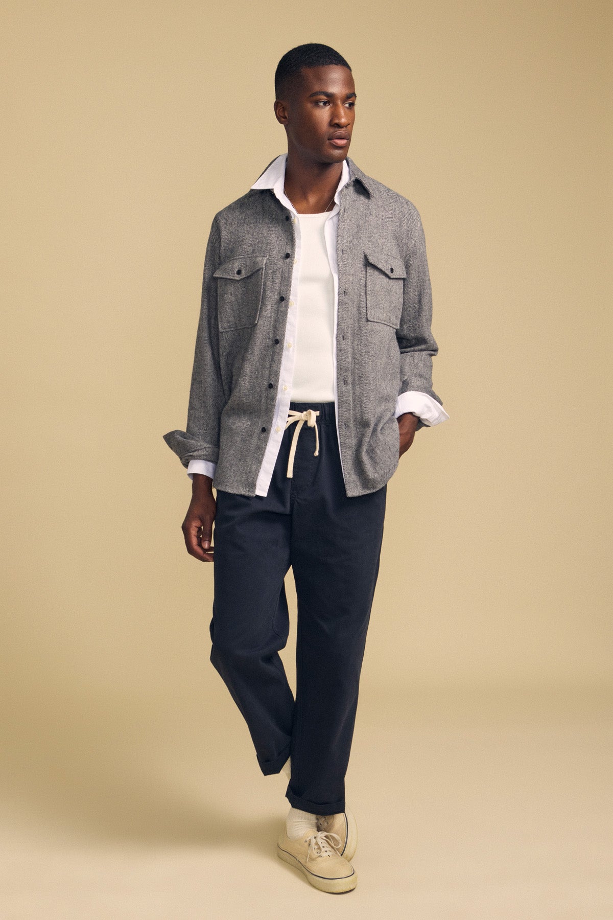 A man stands against a beige background wearing the MAURICE SHIRT JACKET by Velvet by Graham & Spencer layered over a white top, paired with dark drawstring pants and beige sneakers.