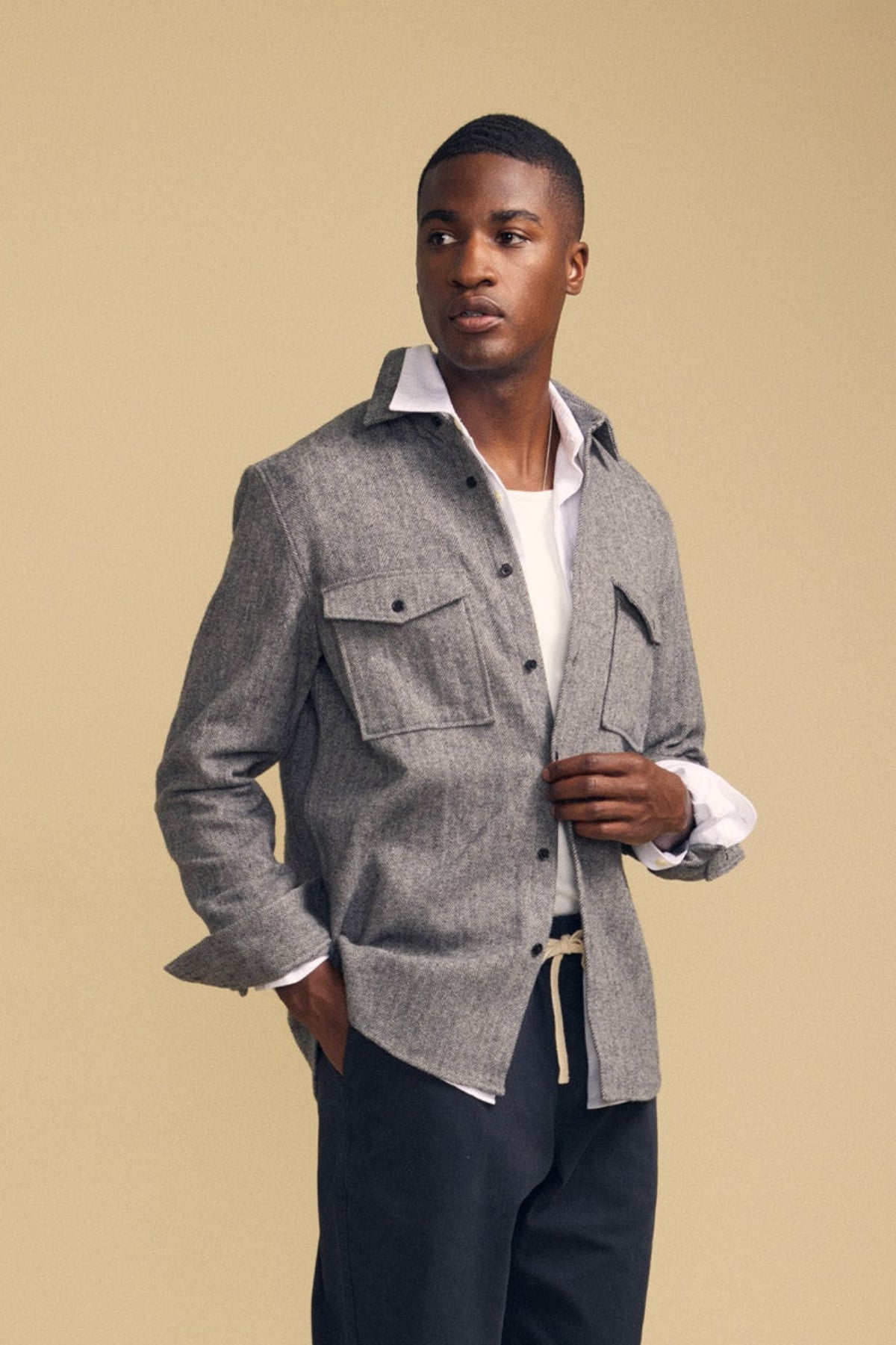 A man wearing the MAURICE SHIRT JACKET by Velvet by Graham & Spencer—a grey button-up overshirt with flap pockets—layered over a white shirt and paired with dark pants stands against a plain beige background.
