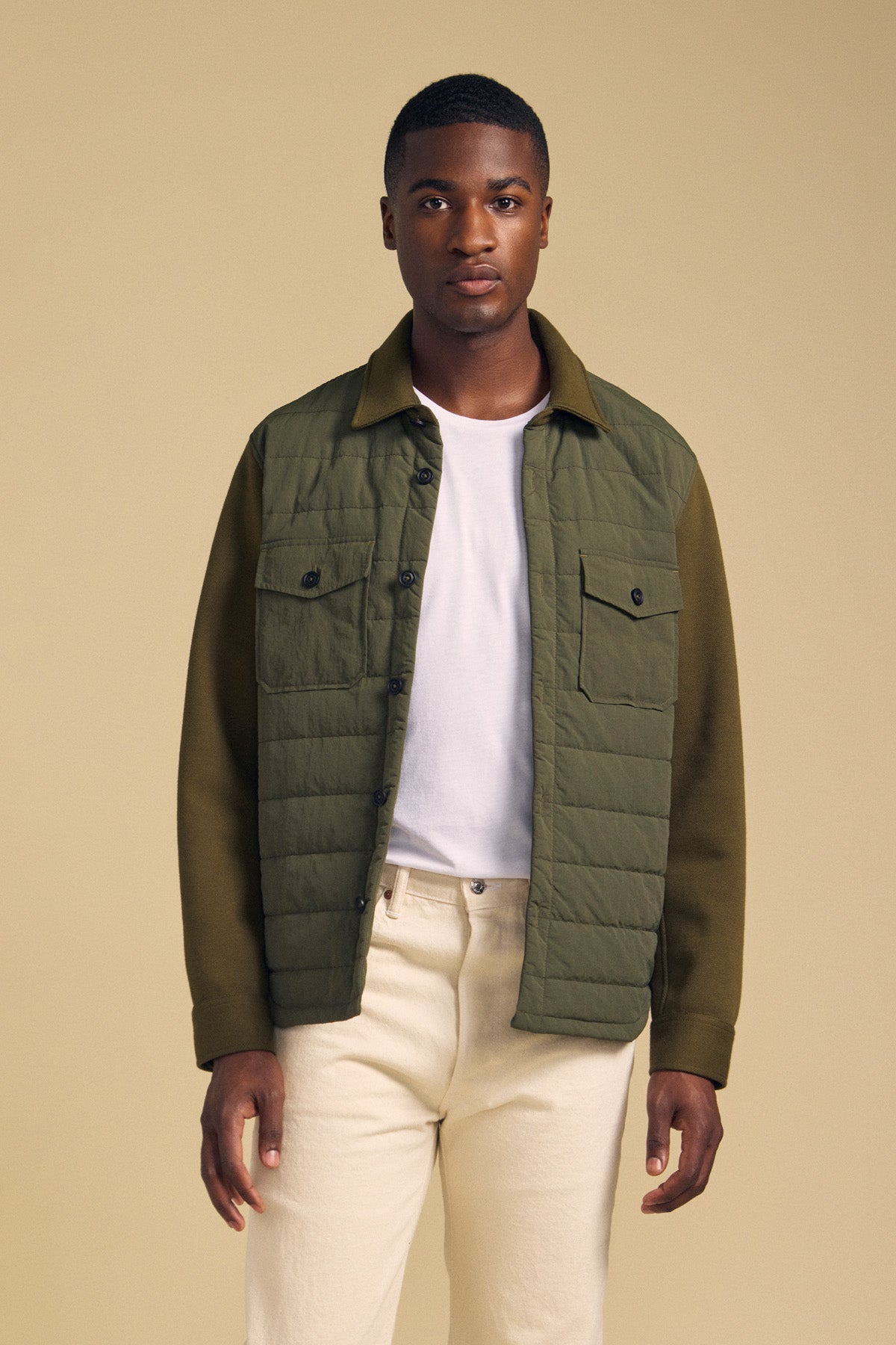 A man wears a Velvet by Graham & Spencer RYKER QUILTED JACKET in olive green over a white t-shirt and cream-colored pants.