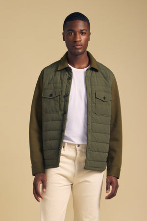 A man wears a Velvet by Graham & Spencer RYKER QUILTED JACKET in olive green over a white t-shirt and cream-colored pants.