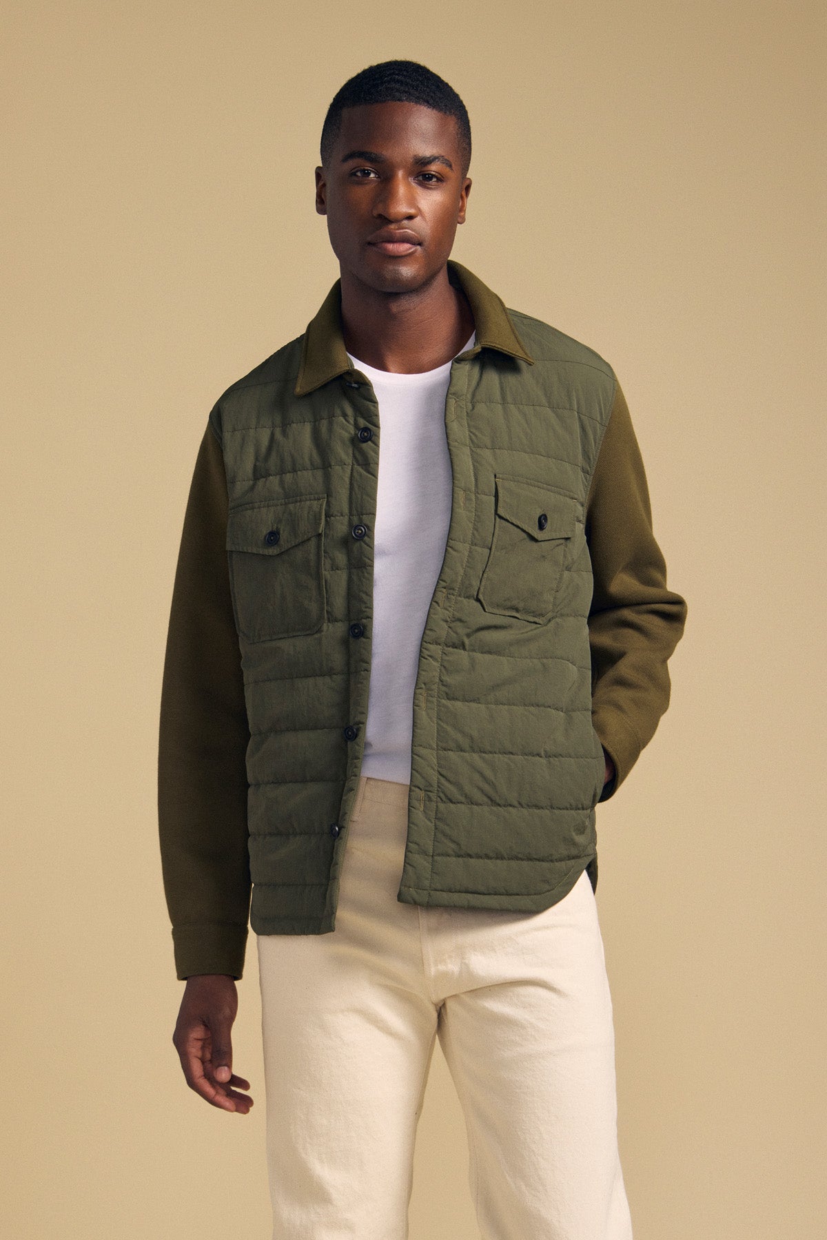 A man wears a Velvet by Graham & Spencer RYKER QUILTED JACKET in olive green over a white t-shirt and cream pants, perfect for transitional weather.-46288448127169