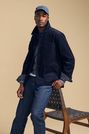 A man wearing the SEGAL CORDUROY JACKET by Velvet by Graham & Spencer, paired with a gray shirt, jeans, and a blue cap, sits on the edge of a wooden chair against a beige background.