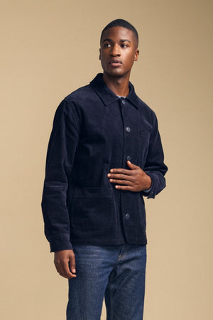 A man wearing the SEGAL CORDUROY JACKET by Velvet by Graham & Spencer and jeans stands against a plain beige background, looking slightly to the side.
