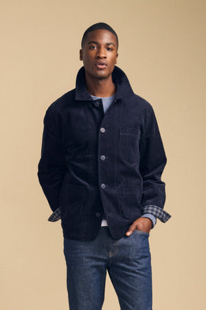 A man models the SEGAL CORDUROY JACKET by Velvet by Graham & Spencer, layered over a gray shirt and blue jeans. He stands against a beige background, looking at the camera with his hands behind his back.