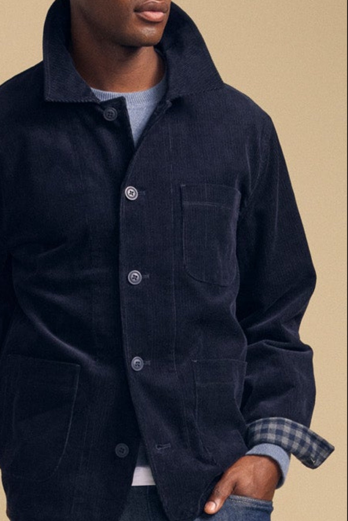 A man wearing the SEGAL CORDUROY JACKET by Velvet by Graham & Spencer in dark blue over a light gray sweater, hand in his pocket, stands against a beige background.