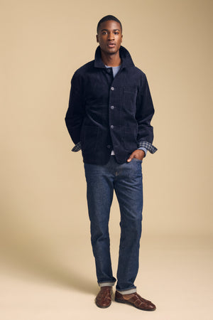 A man stands against a beige background wearing the SEGAL CORDUROY JACKET by Velvet by Graham & Spencer, paired with a plaid shirt, blue jeans, and brown sandals, with his hands in his jacket pockets.