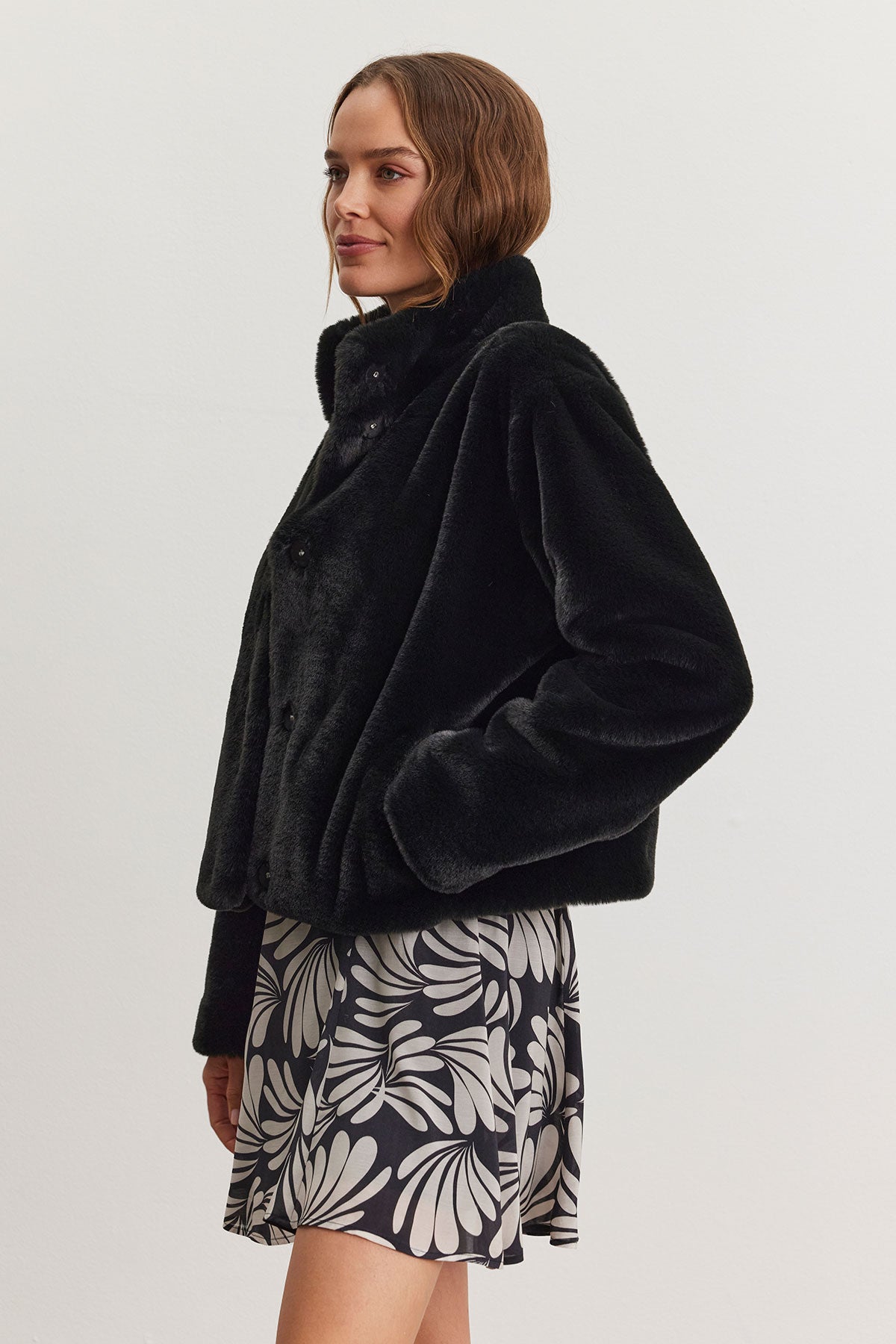 A person modeling the SHEENA LUXE FAUX FUR JACKET by Velvet by Graham & Spencer, featuring a cozy fit and mock neck collar in black, stands against a white background, complemented by a patterned skirt.-38301434282177