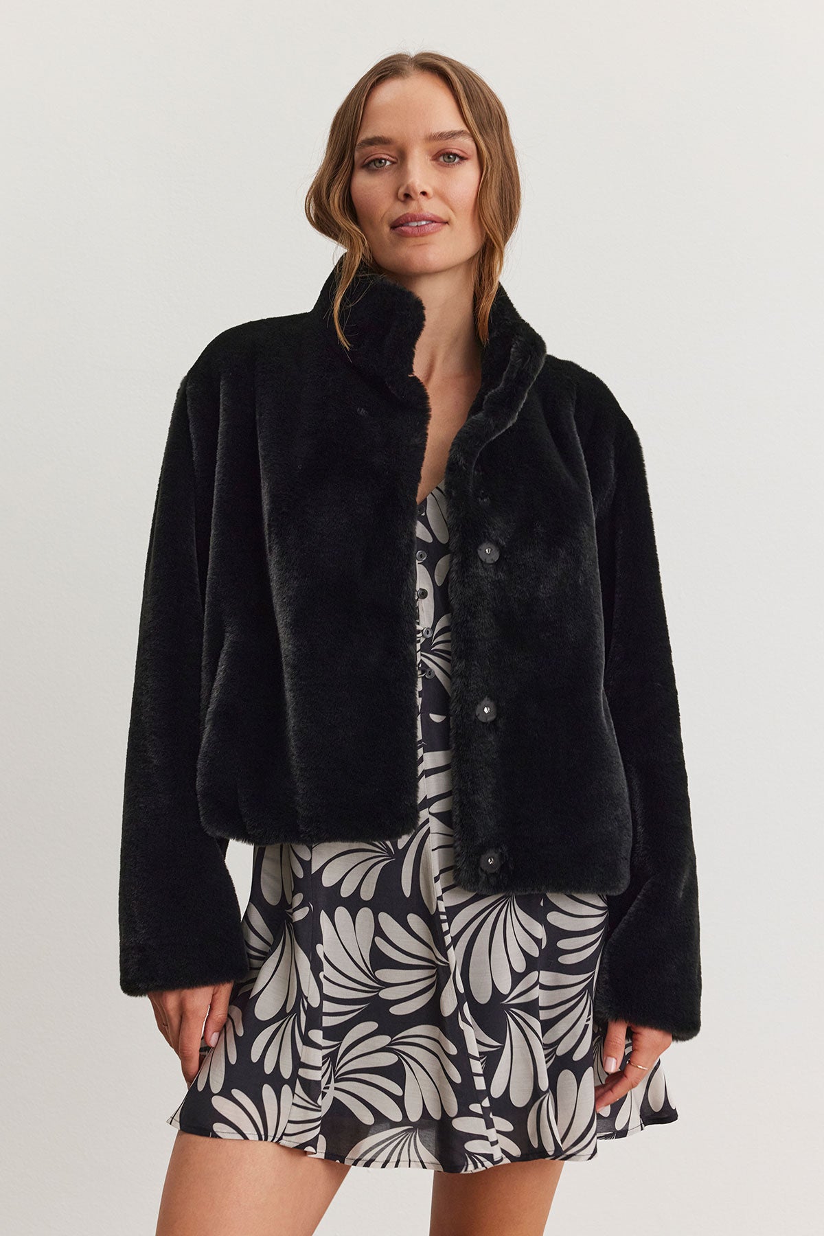   A woman dressed in the SHEENA LUXE FAUX FUR JACKET by Velvet by Graham & Spencer, characterized by its cozy fit, layers it over a black and white floral dress while standing against a plain background. 