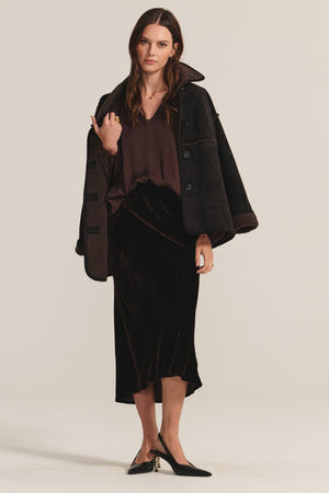 A woman wears a dark shearling coat draped over her shoulders, a brown blouse, and the FELICITY SKIRT by Velvet by Graham & Spencer—black silk velvet with an elastic waistband—paired with black heels featuring gold accents.