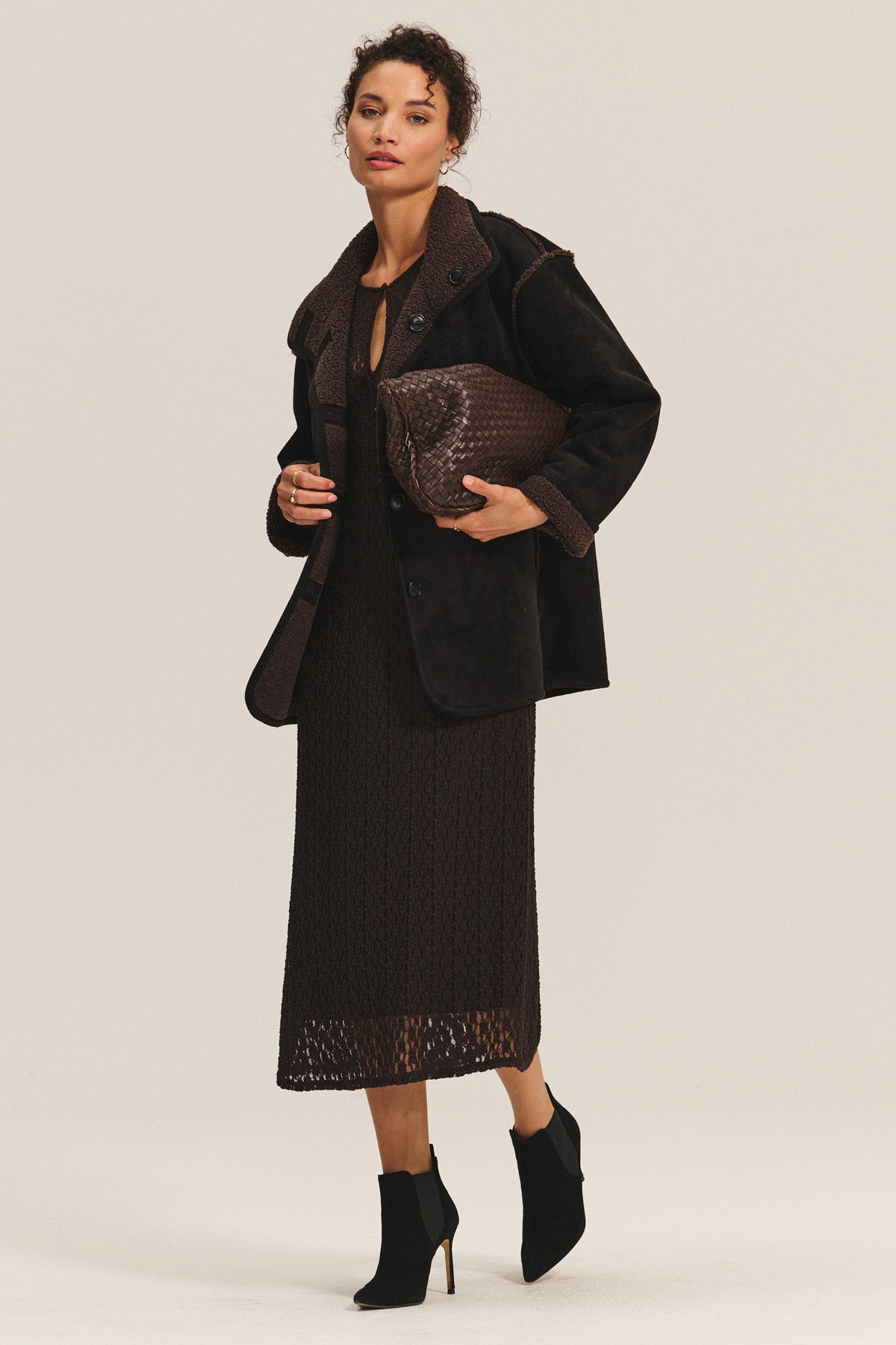 A woman stands against a plain background wearing Velvet by Graham & Spencer's SHONA SHERPA REVERSIBLE JACKET, a dark brown textured dress, black ankle boots, and holding a large woven dark brown clutch bag.-40263669612737