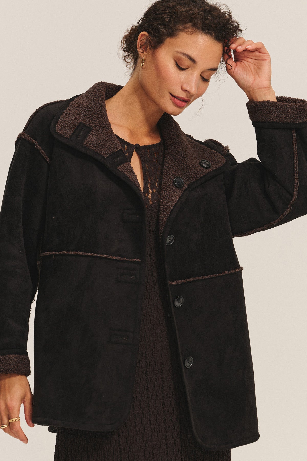 A woman wears the SHONA SHERPA REVERSIBLE JACKET by Velvet by Graham & Spencer over a textured dress, standing against a plain background, looking down with one hand raised to her head.-40263669579969