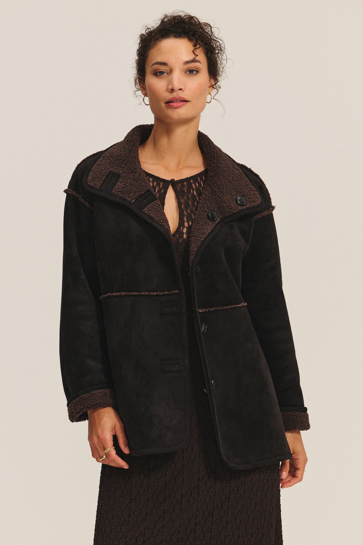 A person stands facing the camera against a plain light background, wearing Velvet by Graham & Spencer’s SHONA SHERPA REVERSIBLE JACKET in dark brown faux suede over a textured brown dress.