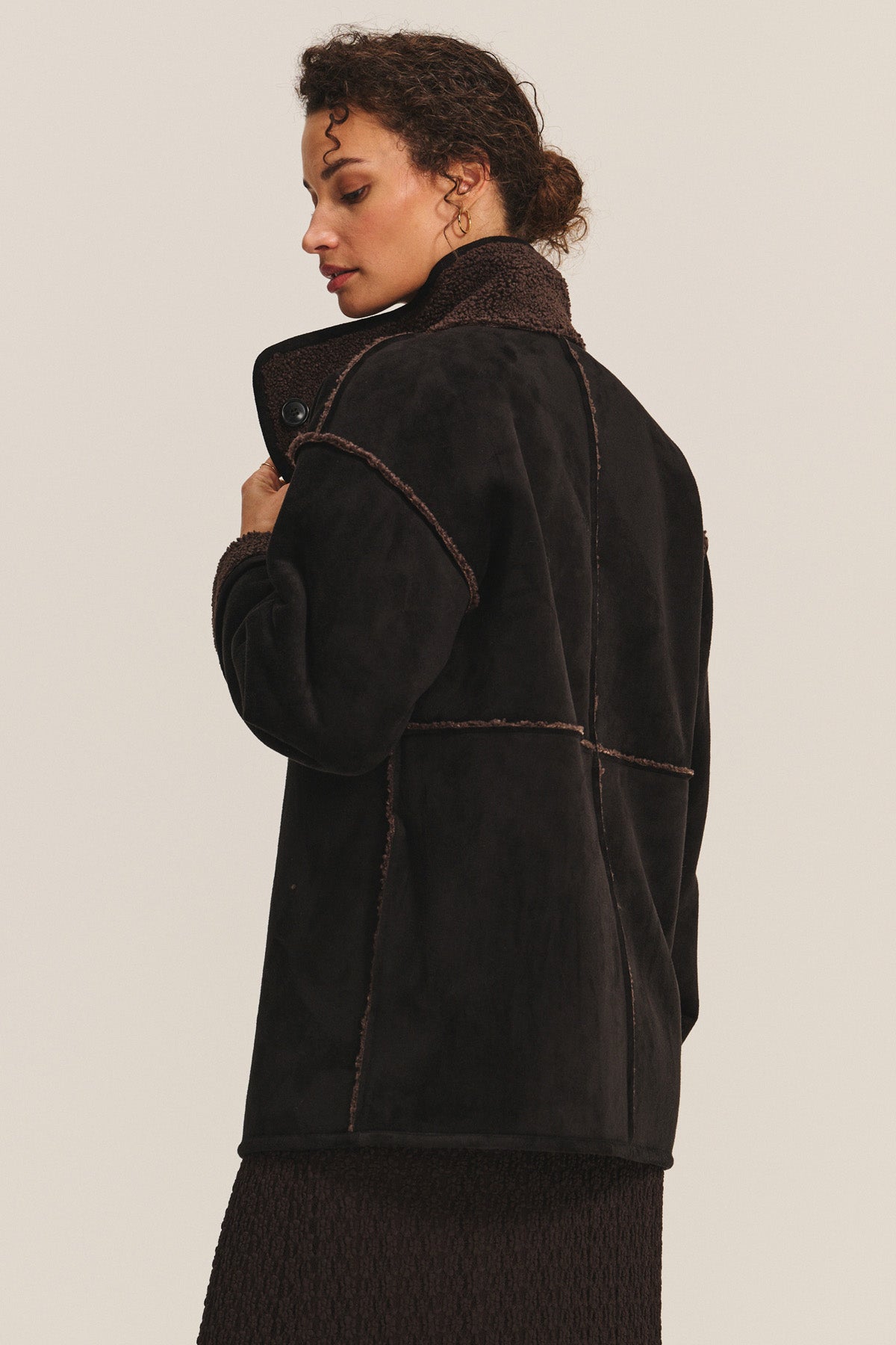 A woman wears the SHONA SHERPA REVERSIBLE JACKET by Velvet by Graham & Spencer—a black faux suede coat with contrast stitching and a high collar—as she stands sideways, looking down against a plain background.