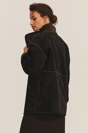 A woman wears the SHONA SHERPA REVERSIBLE JACKET by Velvet by Graham & Spencer—a black faux suede coat with contrast stitching and a high collar—as she stands sideways, looking down against a plain background.