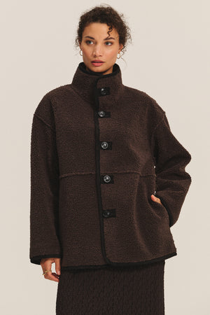 A woman models the Velvet by Graham & Spencer SHONA SHERPA REVERSIBLE JACKET—dark brown, button-up, high-collar, with cozy faux sherpa details—one hand in her pocket, standing against a plain light background.