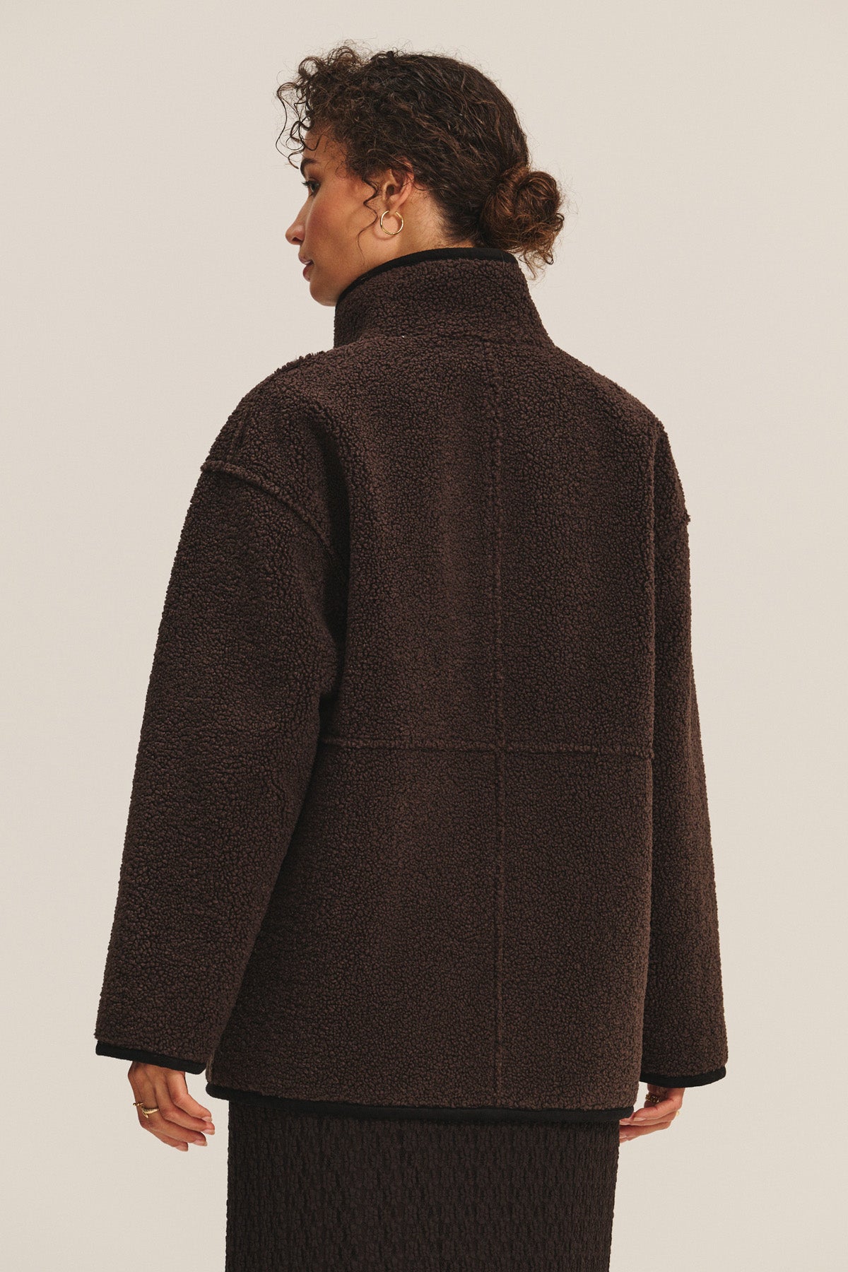 A person stands with their back to the camera, wearing the SHONA SHERPA REVERSIBLE JACKET by Velvet by Graham & Spencer, a dark brown textured, high-collared faux suede piece, paired with a matching skirt against a plain background.