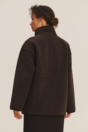 A person stands with their back to the camera, wearing the SHONA SHERPA REVERSIBLE JACKET by Velvet by Graham & Spencer, a dark brown textured, high-collared faux suede piece, paired with a matching skirt against a plain background.