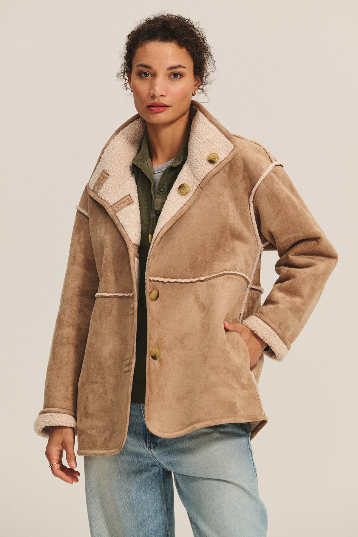 A woman wears the SHONA SHERPA REVERSIBLE JACKET by Velvet by Graham & Spencer in tan faux suede with large buttons, paired with an olive green shirt and light blue jeans, standing against a plain background.