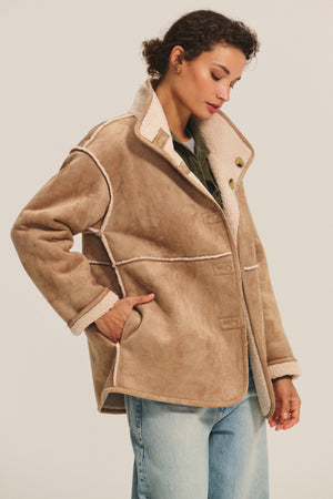 A woman wears the SHONA SHERPA REVERSIBLE JACKET by Velvet by Graham & Spencer—a tan faux suede jacket with white trim—paired with blue jeans, standing against a plain light background.