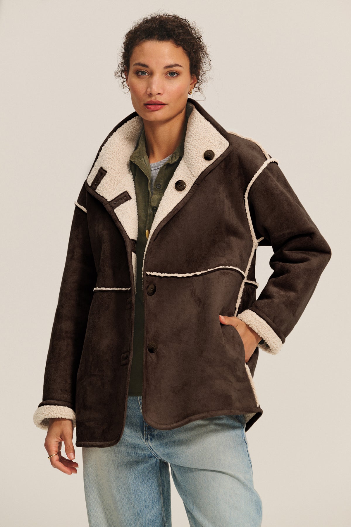   A person wears the SHONA SHERPA REVERSIBLE JACKET by Velvet by Graham & Spencer—brown faux suede with white lining—over a green shirt and light blue jeans, standing against a plain background. 