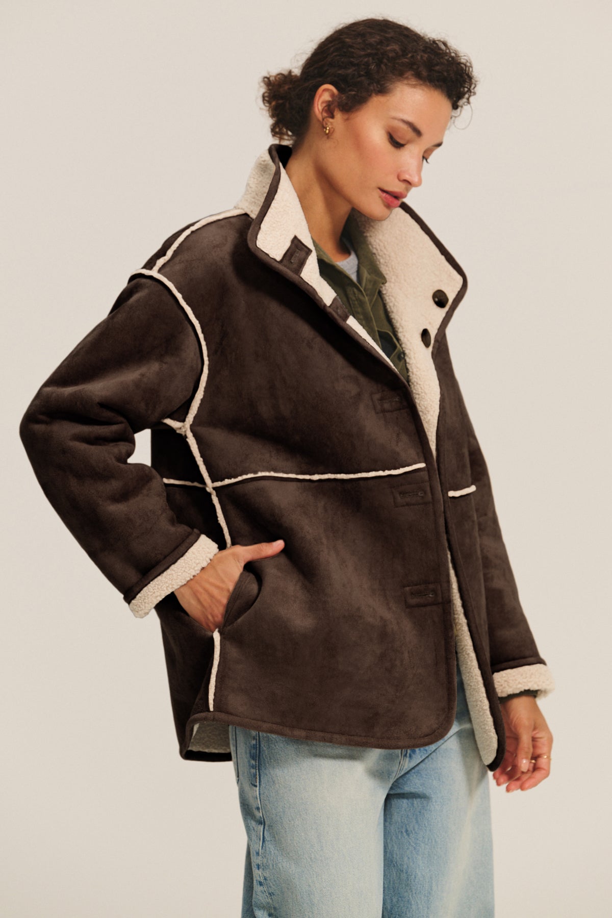   A woman wears the Velvet by Graham & Spencer SHONA SHERPA REVERSIBLE JACKET with cream trim over a green top and light blue jeans, standing against a plain background. 